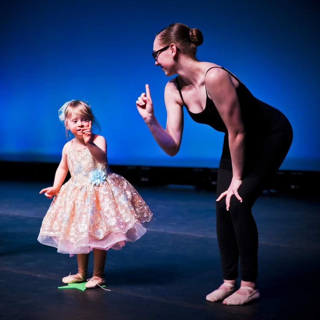 Young girl in a sparkly dress dances on stage with her teacher, who gently guides her with a smile. Both share a joyful moment under bright stage lights, celebrating movement and connection.