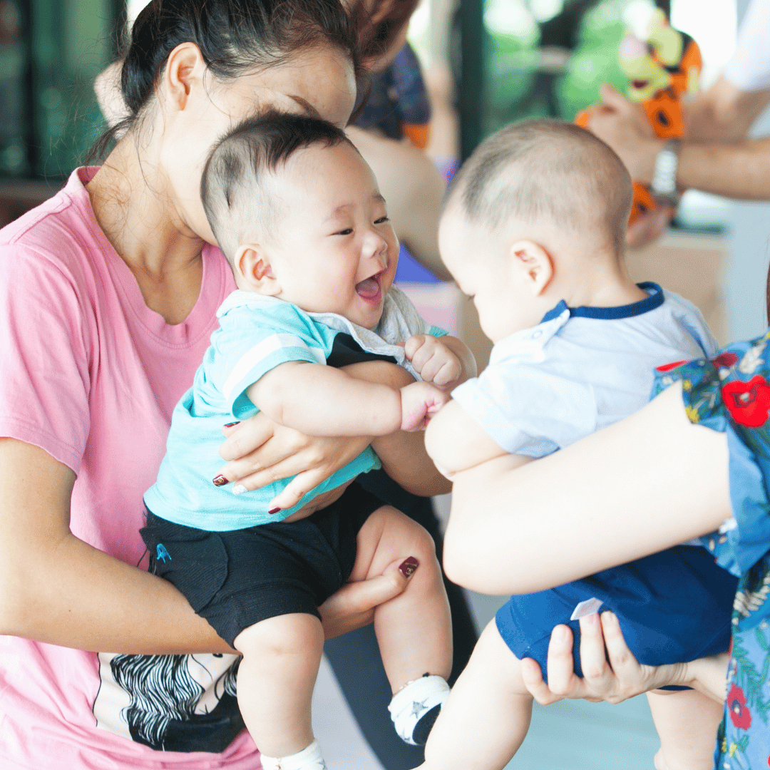 Two moms are holding their babies facing each other and smiling.
