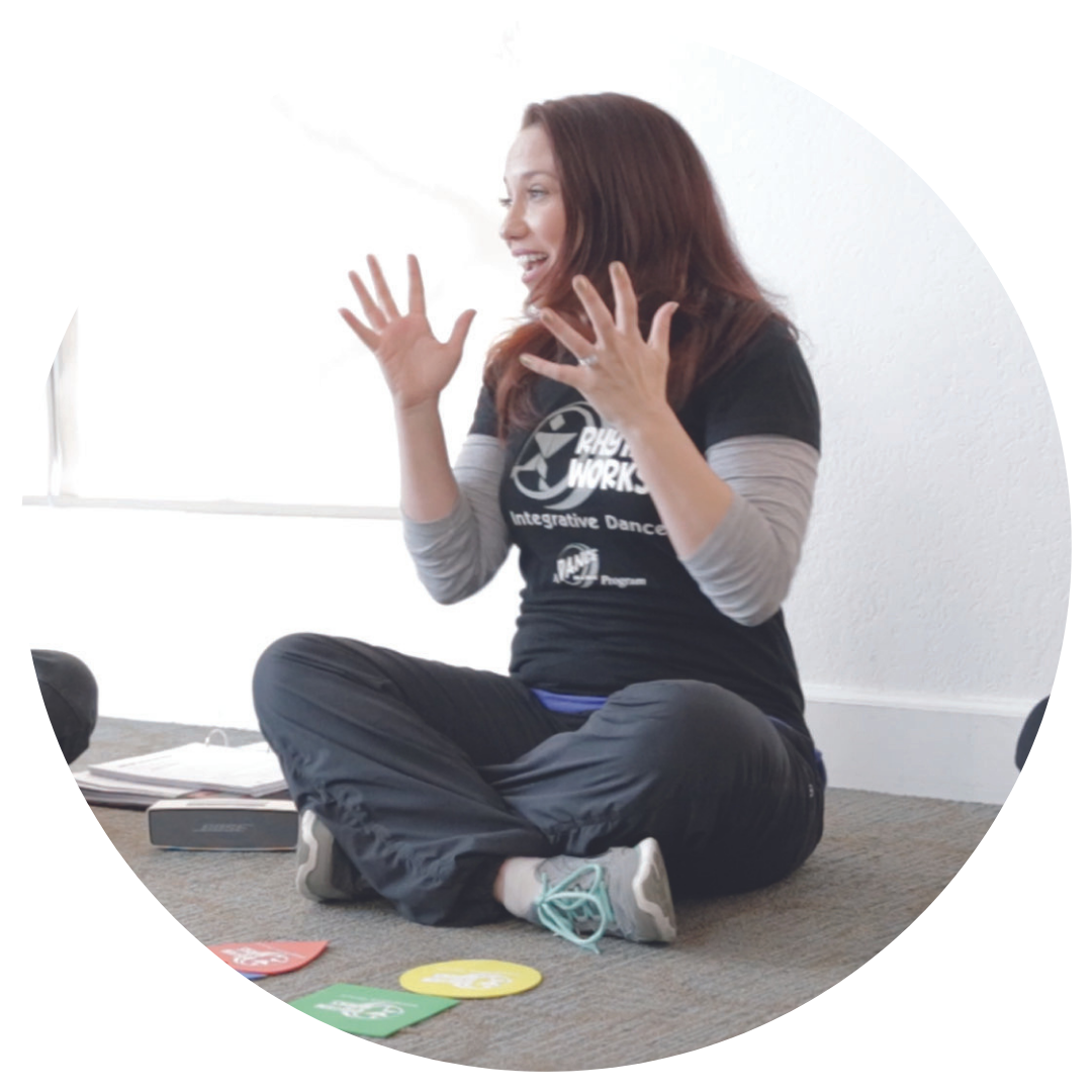 Rhythm Works Integrative Dance instructor sitting cross-legged on the floor, smiling and using expressive hand gestures while teaching. Colorful activity markers are on the floor in front of her.