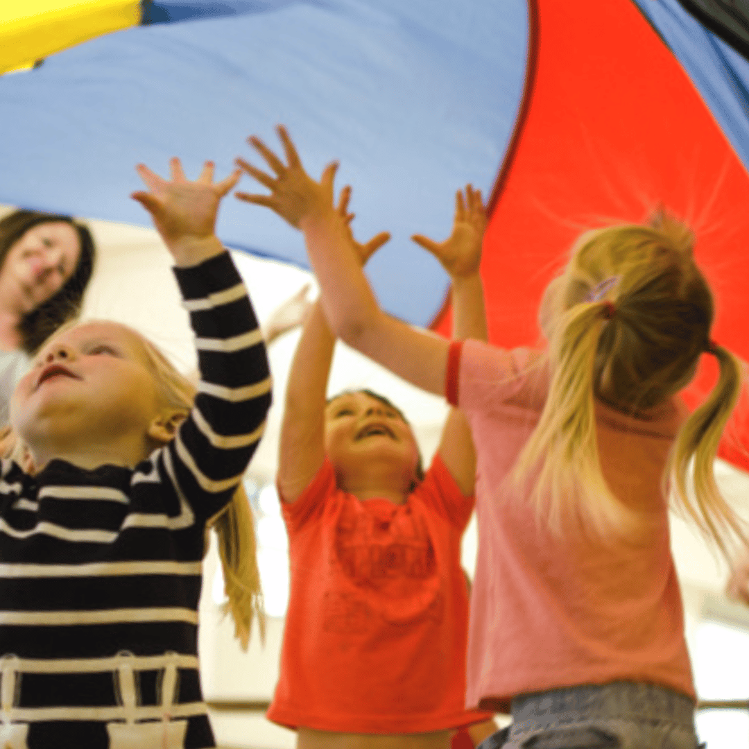 Three preschool aged girls are smiling underneath a colorful parachute.