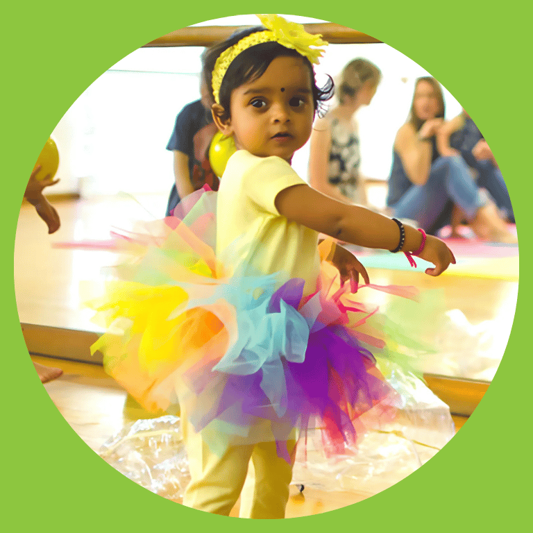 A toddler girl is wearing a yellow shirt and leggings with a rainbow colored tutu and yellow flower in her hair.