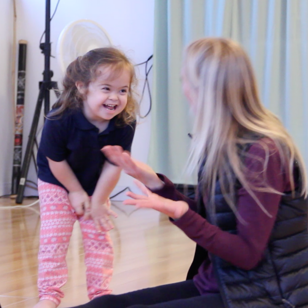 Young girl with a big smile claps hands while interacting with her teacher during an adaptive dance class. Both are engaged and joyful, highlighting connection and fun through movement.