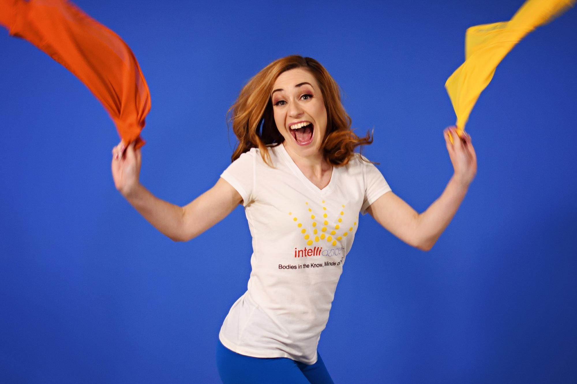 A women wearing a white t-shirt is joyfully jumping while holding onto an orange and a yellow scarf.