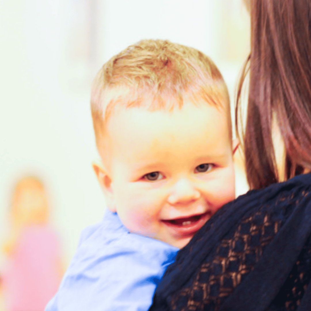 A toddler looking over the shoulder of his mom and smiling.