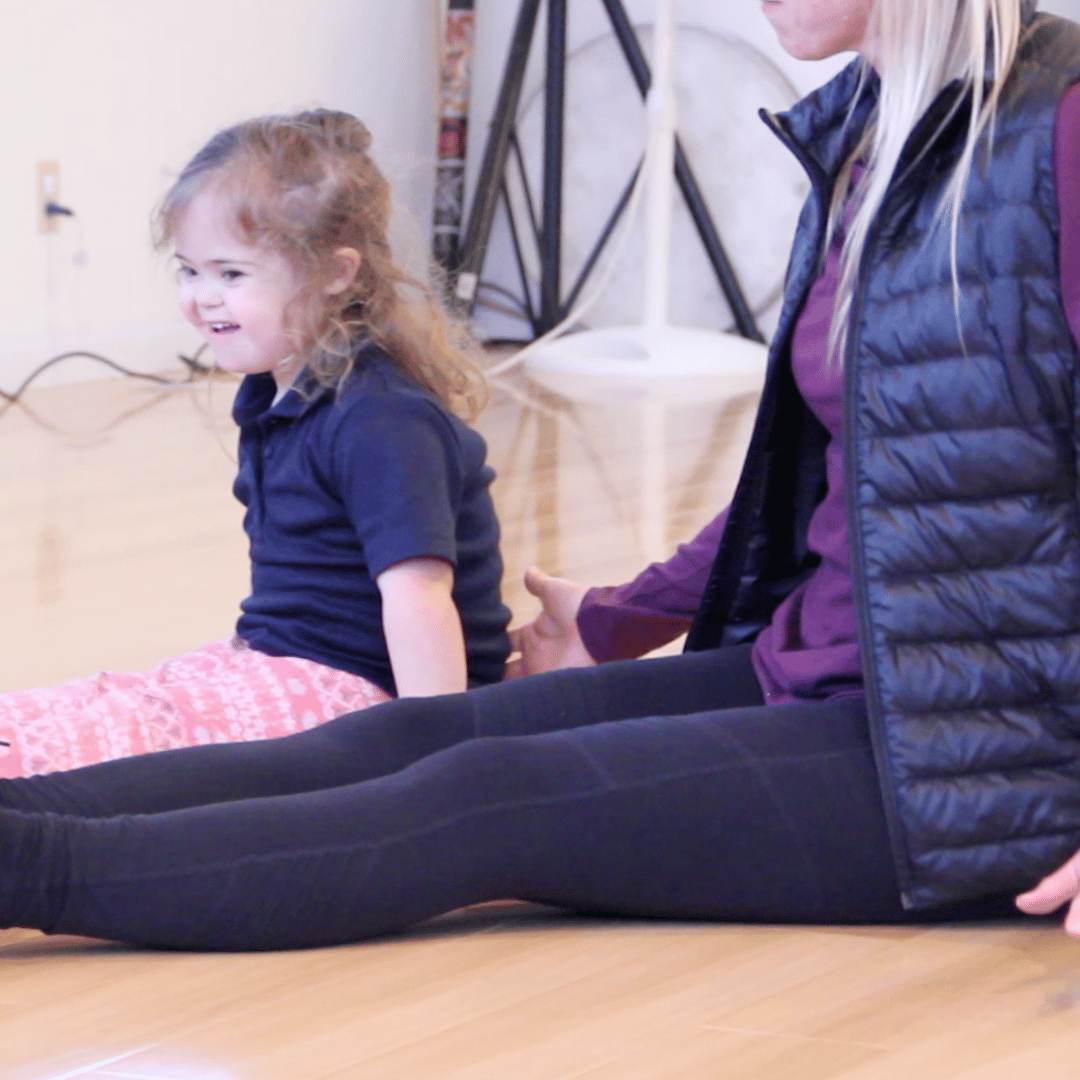 A young dancer is sitting on the floor with her legs stretched out long in front of her. Her Rhythm Works Integrative Dance teacher assistant is sitting next to her with her hand on the student's lower back.