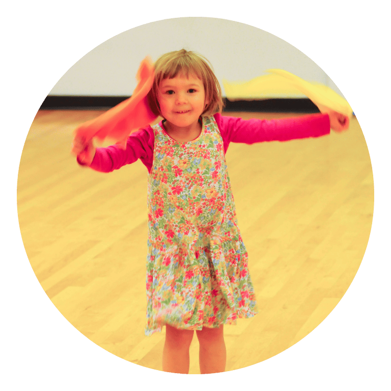 A preschool aged girl is happily dancing while holding two scarves.