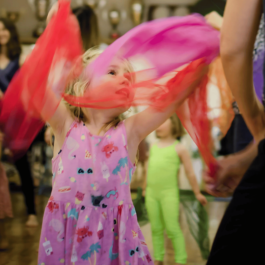 Young girl with blonde hair is wearing a pink dress and dancing with two scarves.