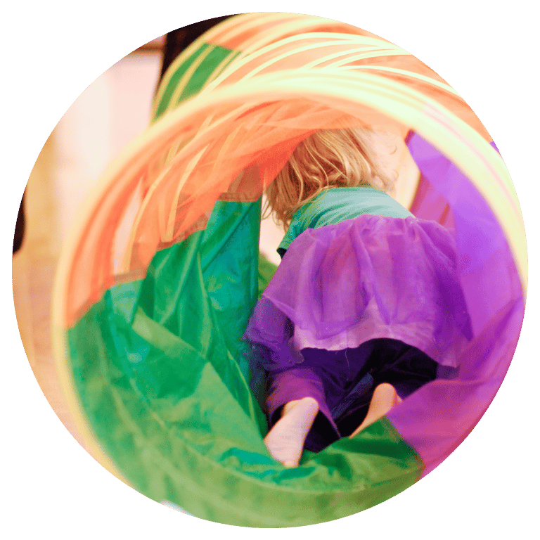 A toddler girl wearing a purple skirt is crawling through a colorful floor tunnel