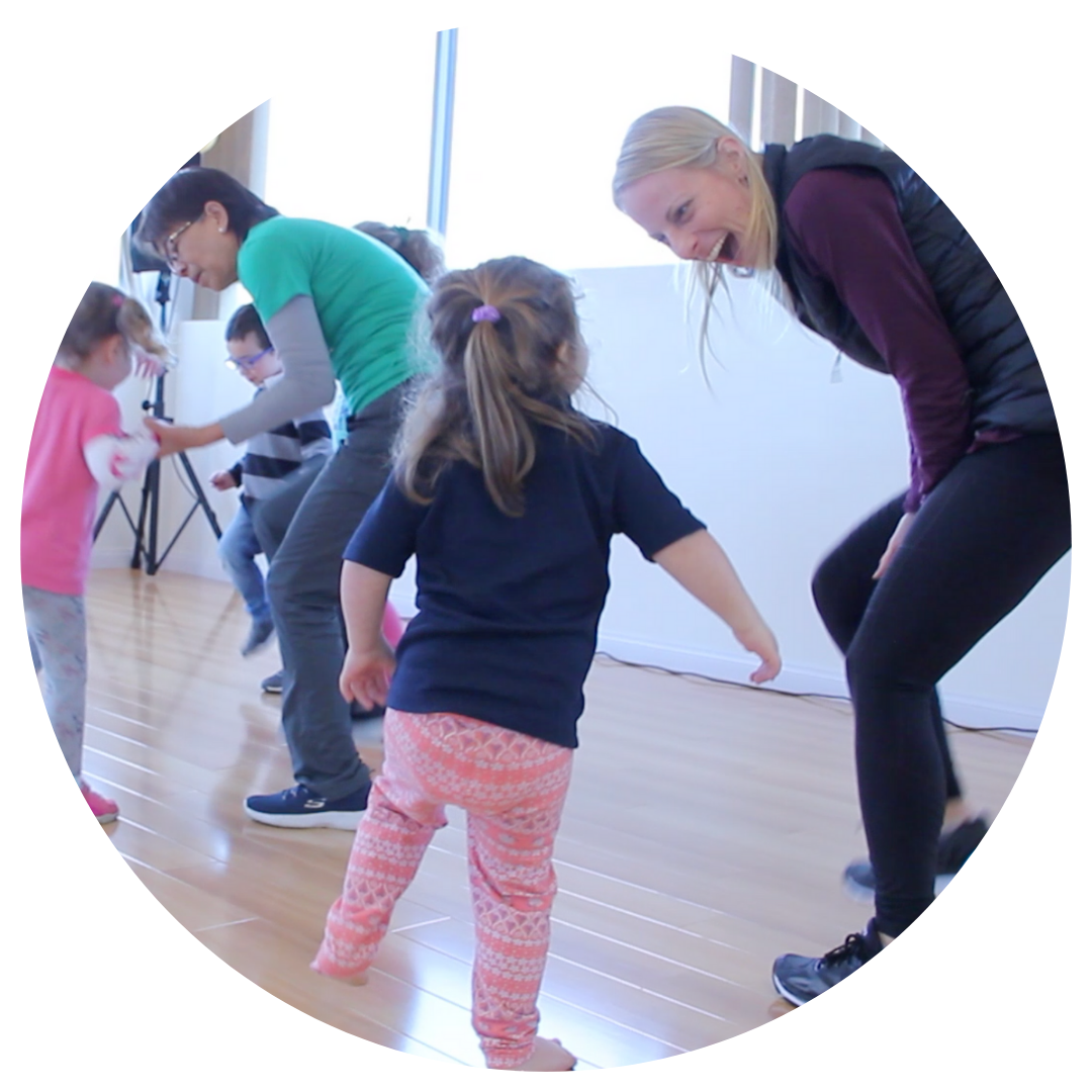 Children and instructors participating in an inclusive dance class. A young girl faces an adult teacher who is smiling and engaging with her, while other children and adults dance together in the background.