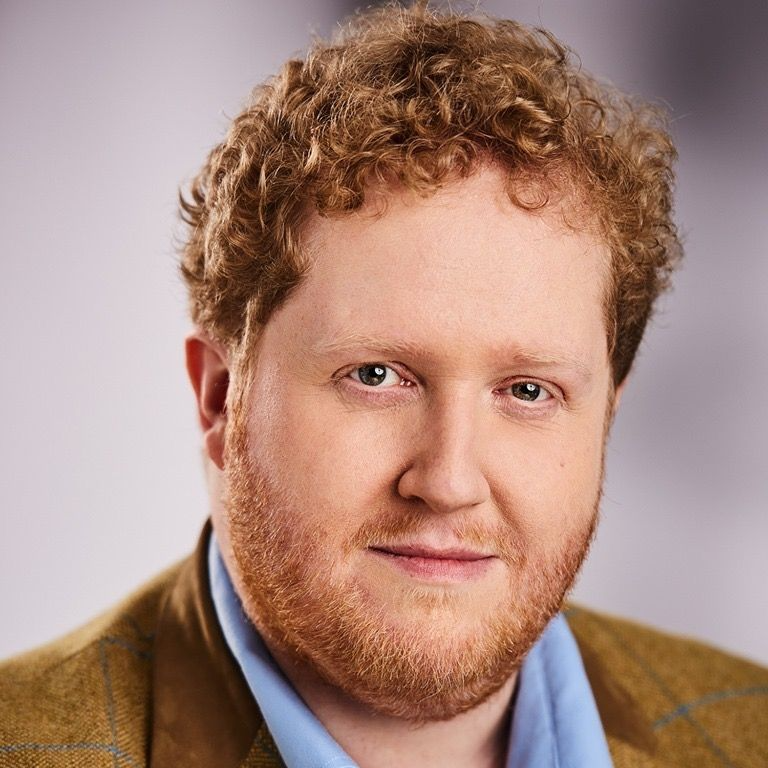 Paddy Cooper, A blonde curly haired guy wearing a brown coat with blue polo in the inside