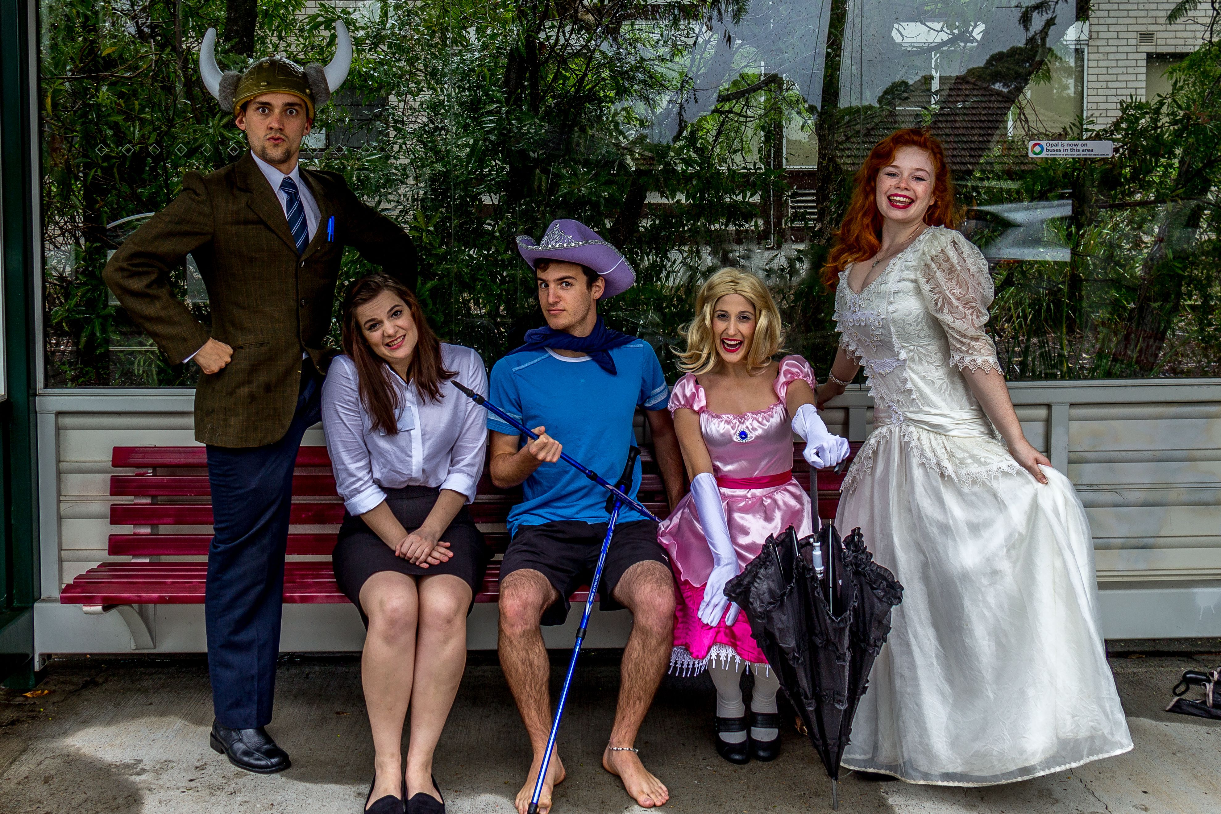Three costumed actors sitting on a red bench with one male wearing a coat and a barbarian helm on the left and a ginger female wearing a wedding dress on the right