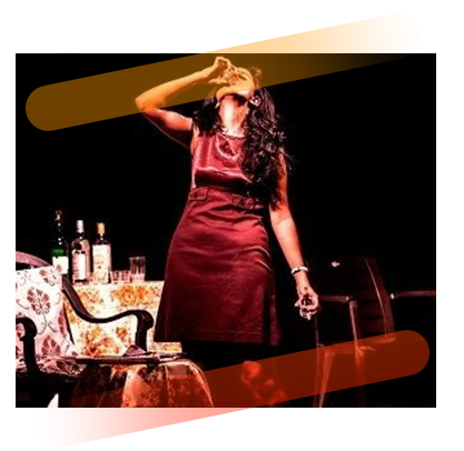 A female on stage wearing a red dress and drinking a glass of liquor with table and chair full of bottles in the background
