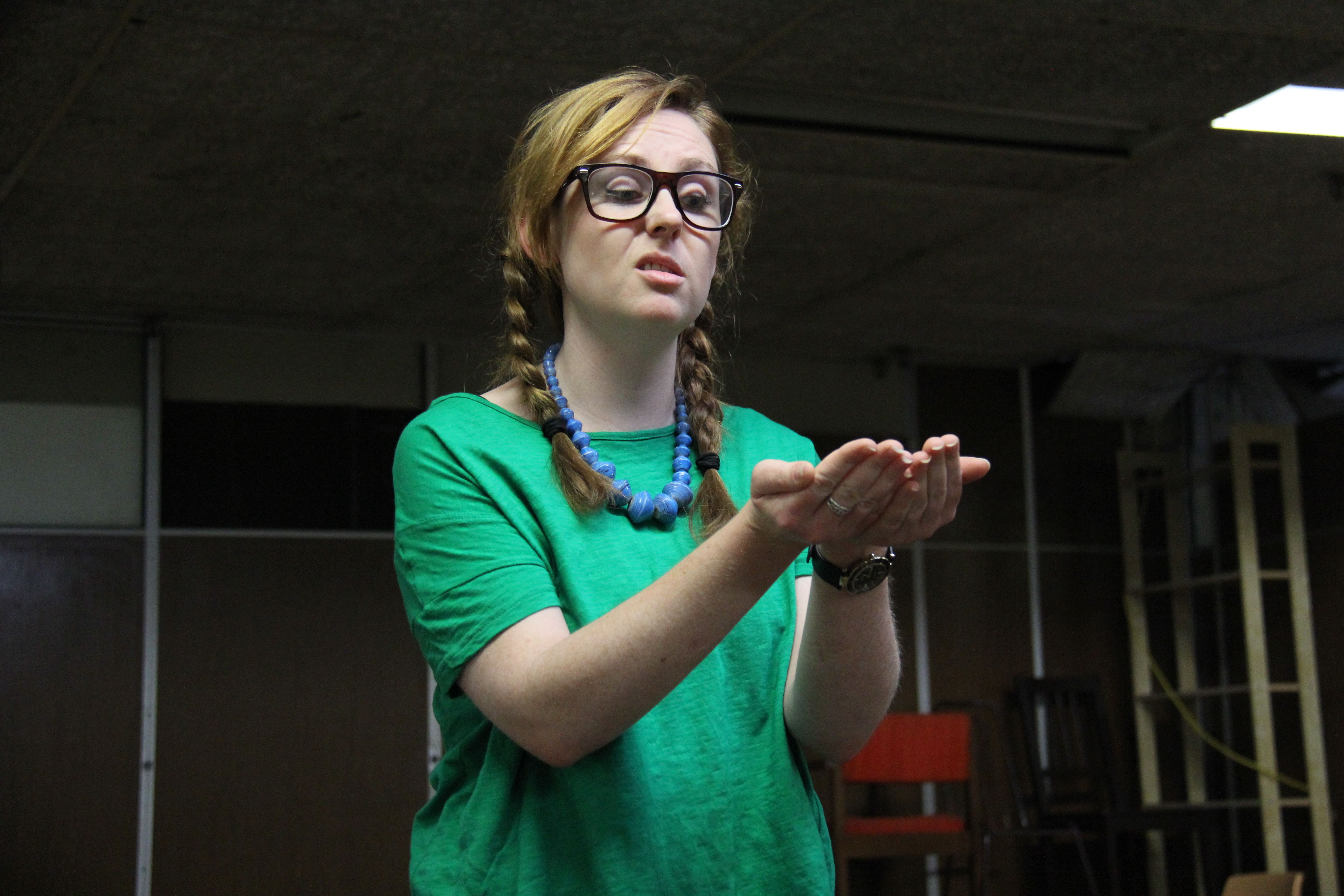 A blonde girl with a pigtail wearing a glasses, green shirt and a blue necklace.