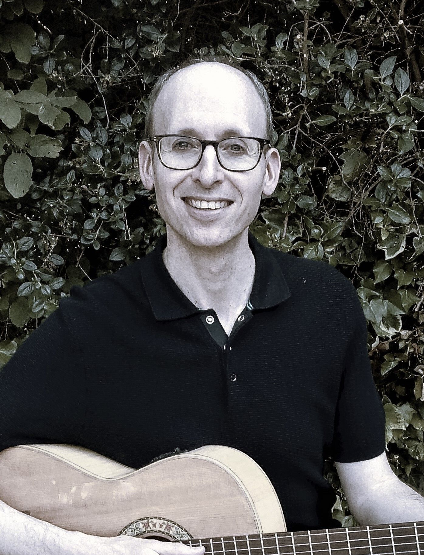 Marc Harris - Smiling white guy wearing a black polo shirt while holding a guitar in a background full of leaves