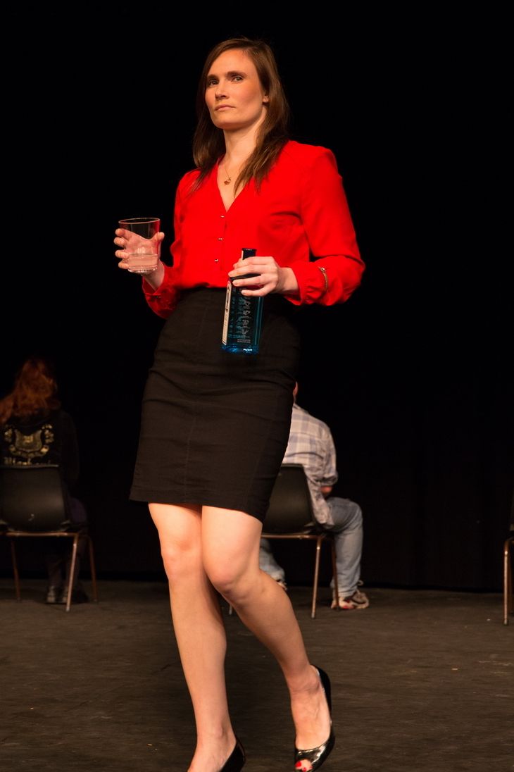 Female actor on stage wearing red blouse and black skirt holding a bottle of liquor and a glass