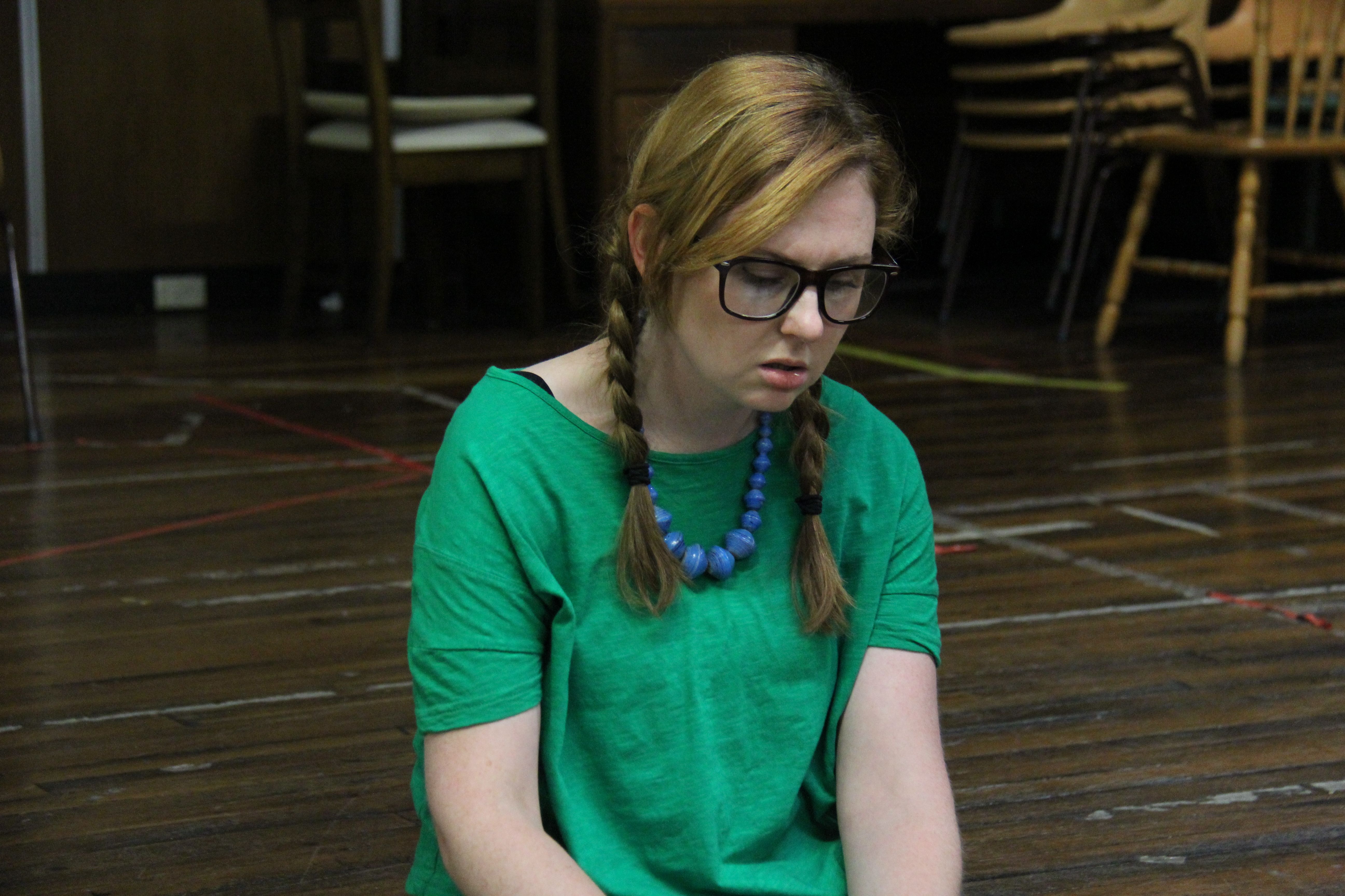 A sad blonde girl with a pigtail wearing a glasses, green shirt and a blue necklace on stage