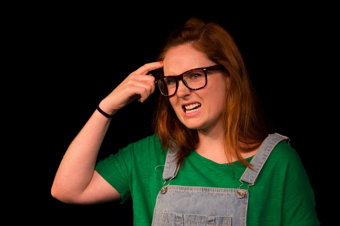 An acting blonde girl wearing a glasses, green shirt with a jumper.