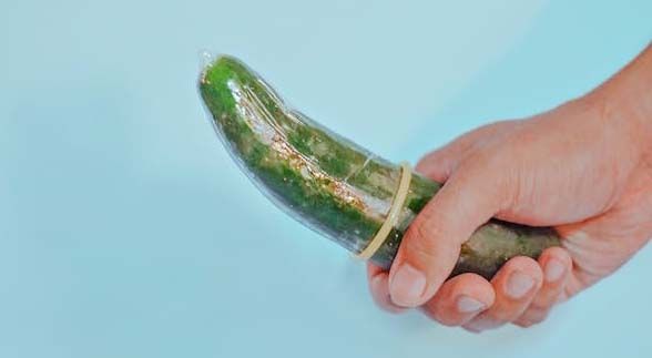 A hand holding a cucumber with condom on it in a bright blue background