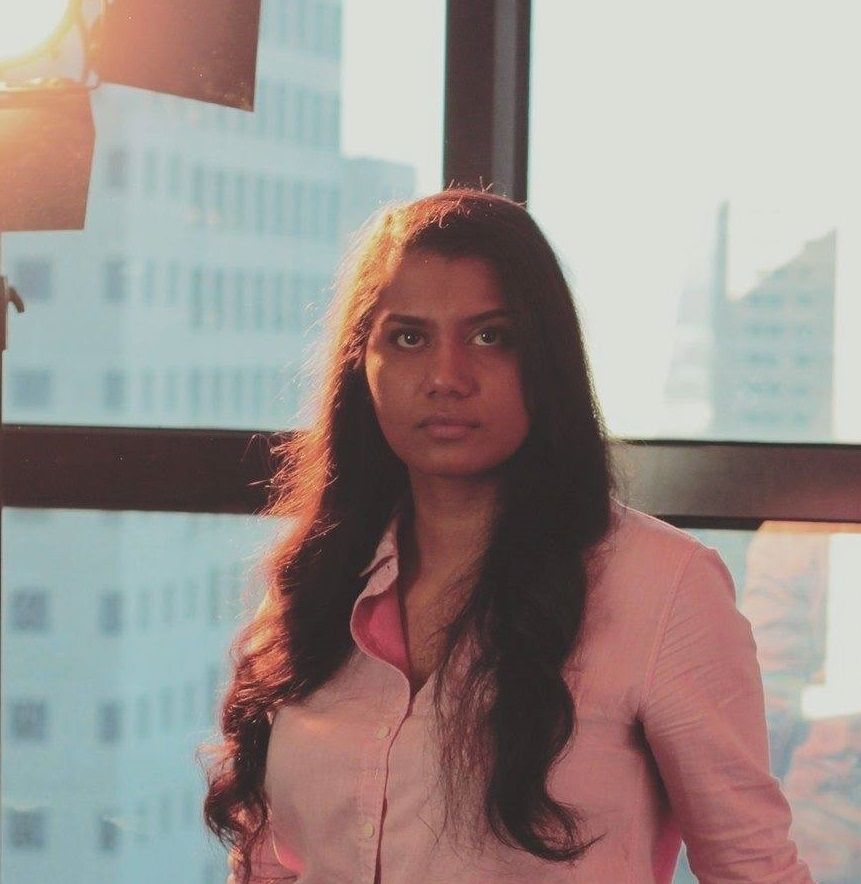 Patsy Ferrer - A woman in a pink shirt standing in front of a window, gazing outside with a serene expression.