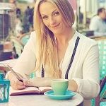 Angharad Thompson Rees, a smiling blonde girl wearing a white formal dress while writing in a notebook in a cafe