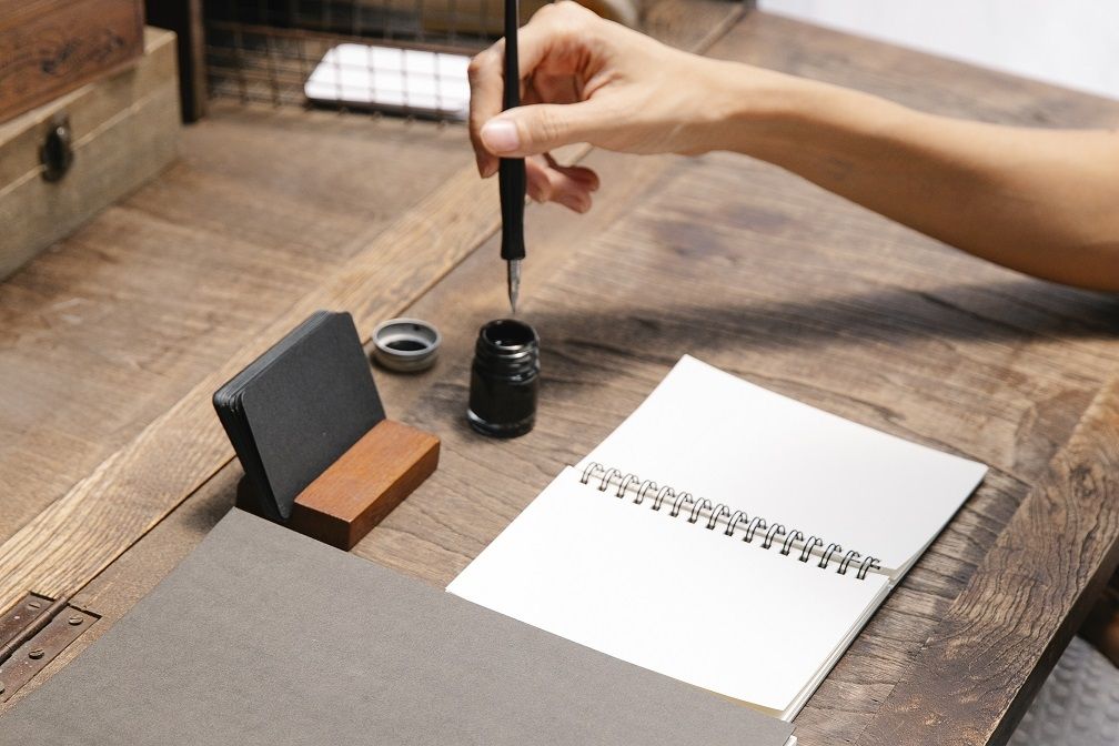 A Hand lying on a wooden table holding a pen that is about to be dipped into ink and write on a piece of paper