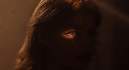 A woman staring at a hole in the wall with her eyes being visible because of the ray of light