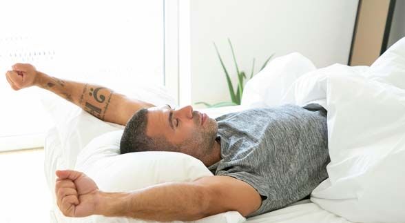 A tattooed man waking up in bed while stretching both of his arms