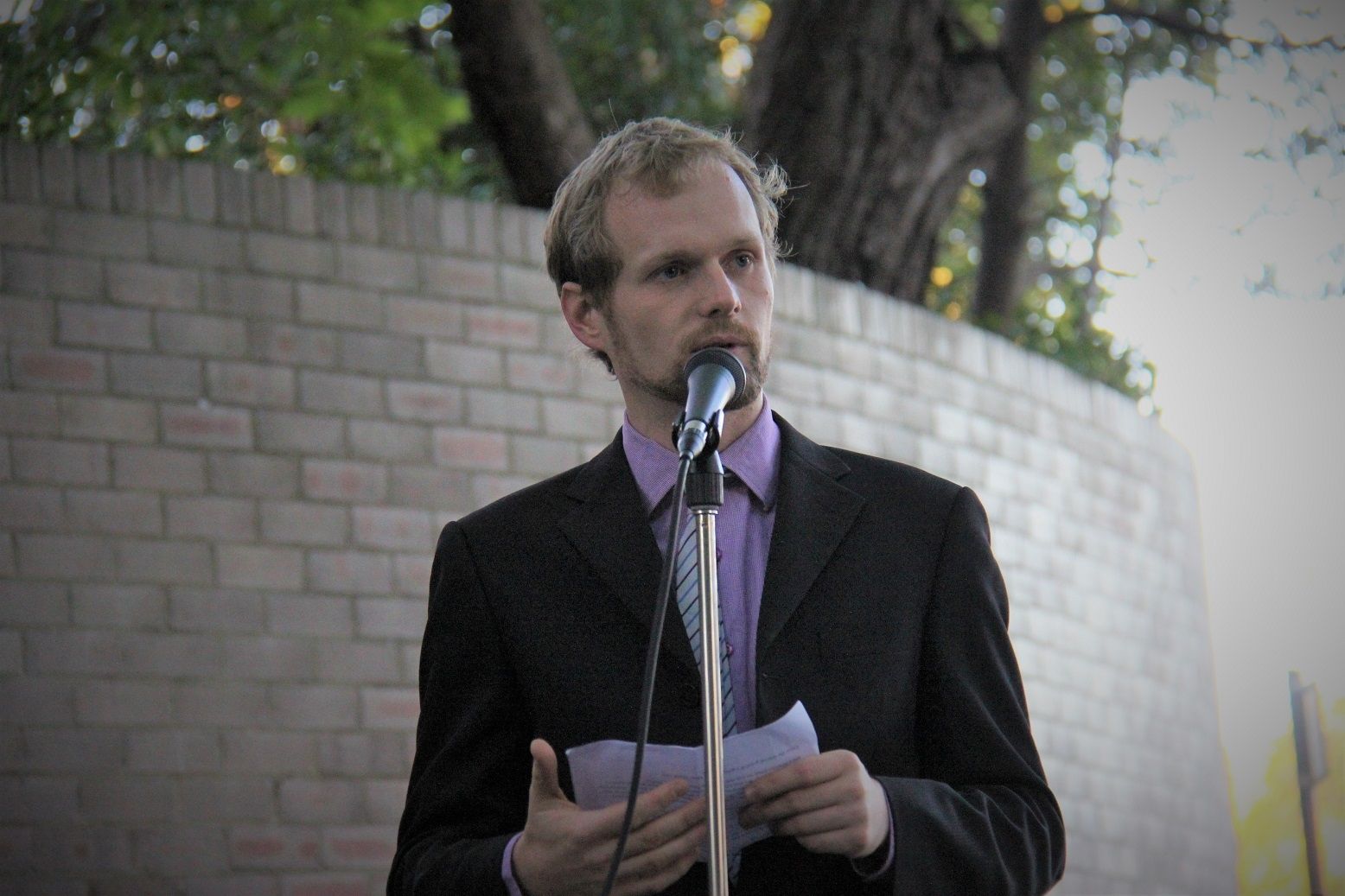 Pete Malicki wearing a purple polo and a coat facing the audience reading a script on a microphone