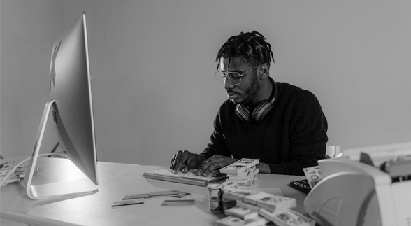 A black and white image of a male in a dreadlocks sitting infront of a desk facing a computer monitor