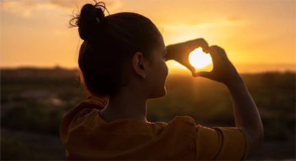 A silhouette of a girl doing a heart sign with her hands in front of the sun