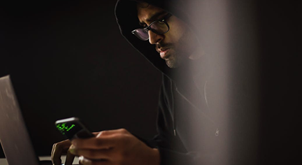 A man in a black hoodie staring at his phone and laptop