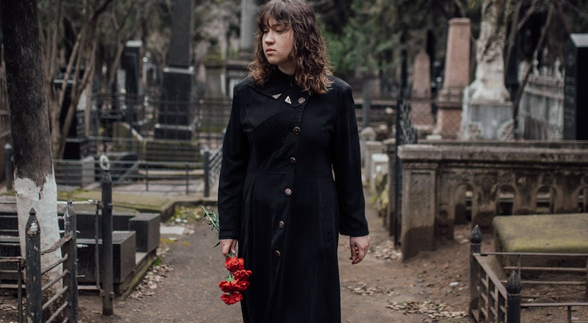 A curly haired female wearing a black dress carrying a red roses at a cemetery