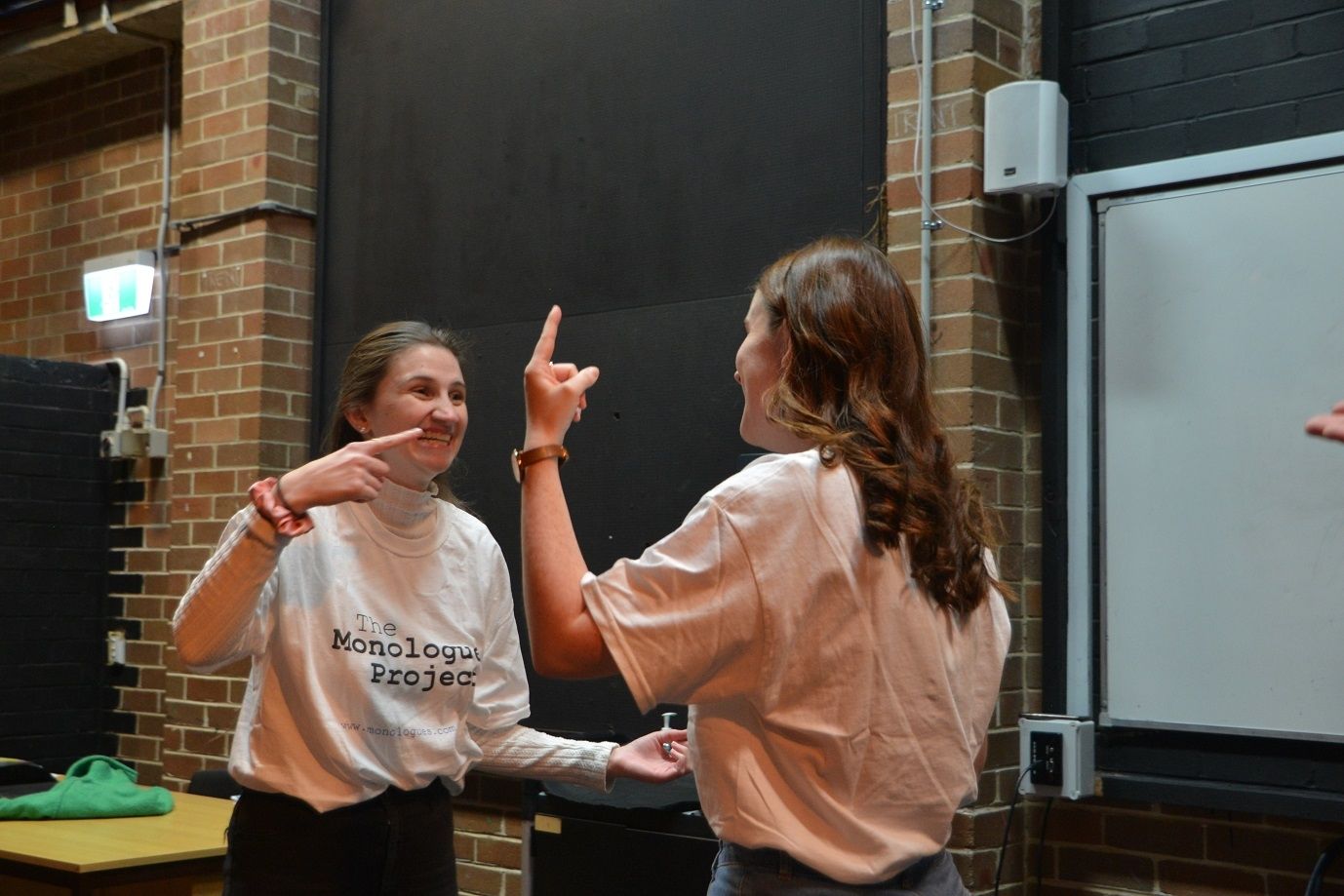 Two female inside an acting office facing each other one point to the other and one pointing upwards