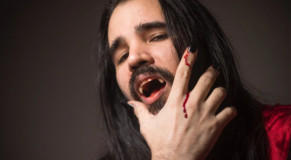 A long haired male with a moustache, a vampire's teeth and a bit of blood dripping in his hands
