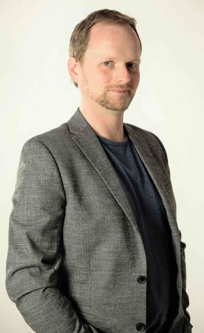 A Portrait Of Pete Malicki in a serious face wearing a gray coat and a navy blue shirt