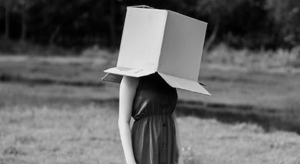 Black and white image of a female in a grass field, cover her head with a box.