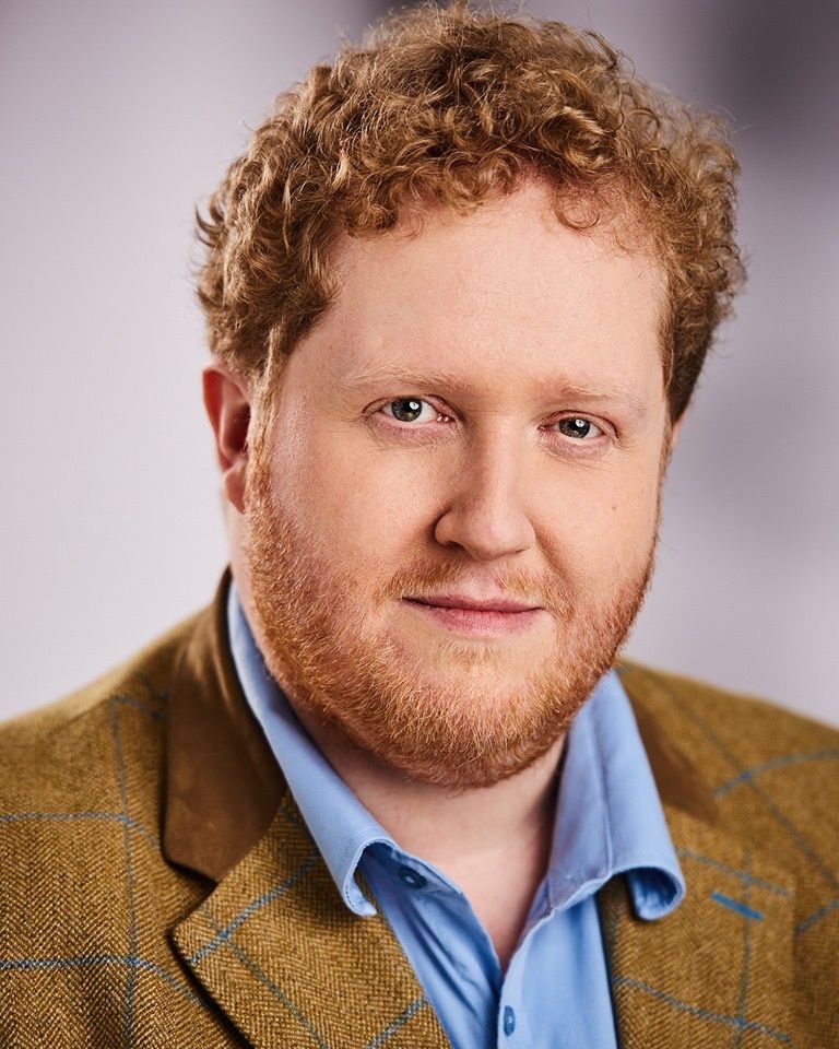 Paddy Cooper, A blonde curly haired guy wearing a brown coat with blue polo in the inside