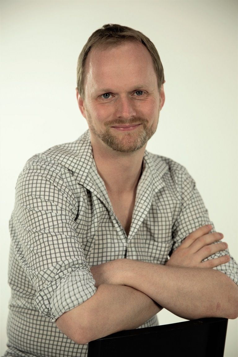 A Portrait of Pete Malicki wearing a checkered white polo leaning his hands on the back of the chair