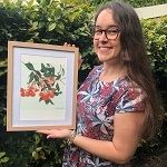 Jessica Harwood - A woman with a curly long hair, smiling, wearing eye glass and a textured dress while holding a painting