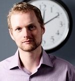 Portrait of Pete Malicki wearing a purple polo with a clock in the background