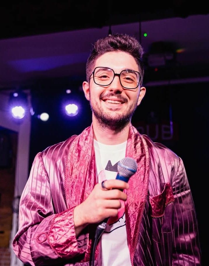 Mike Savuica - A cool bearded guy wearing a glasses, pink coat and white shirt in the inside while holding a microphone