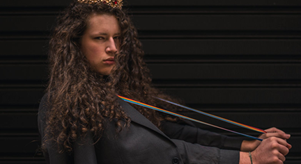 A long curly haired guy with a crown on his head while pulling a rubber band attached to his neck