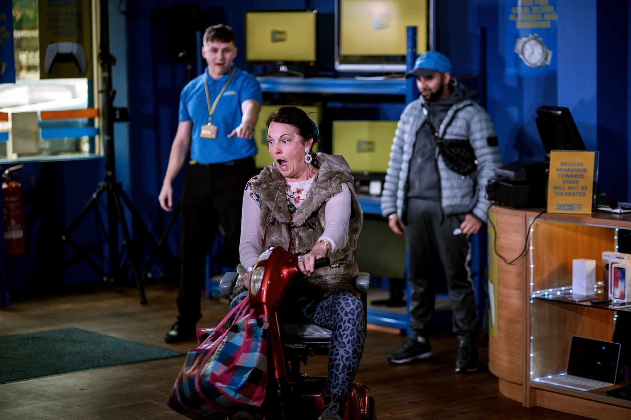 A middle aged woman wearing a pink blouse and a leopard printed leggings while riding an electric wheelchair with two guys in the background
