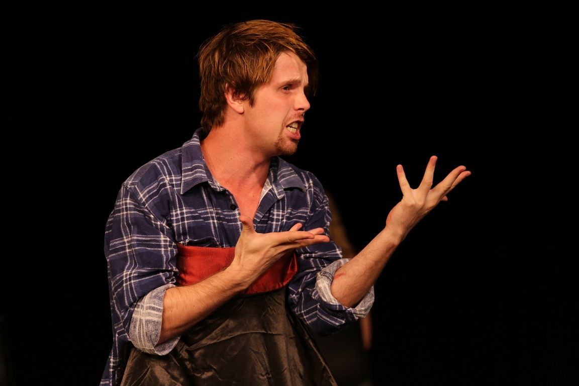 Male actor doing hand gesture wearing blue checkered polo on a black background