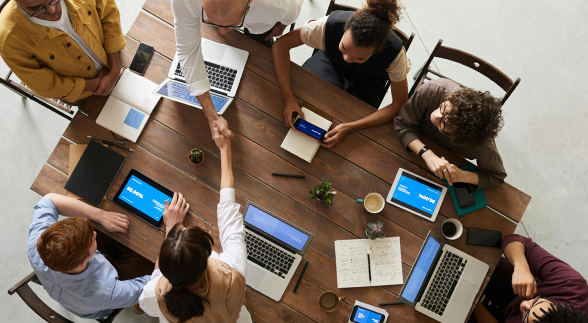 A group of people in an office meeting sitting down in a steel chair and wooden table with laptops in the middle