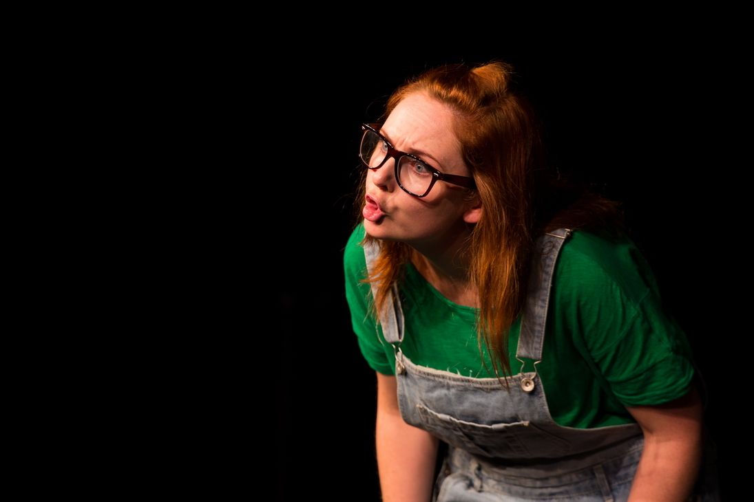 An acting blonde girl with a pigtail wearing a glasses, green shirt with a jumper.