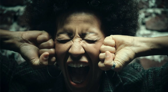 A curly haired female clenching both of her fists to her cheeks while shouting