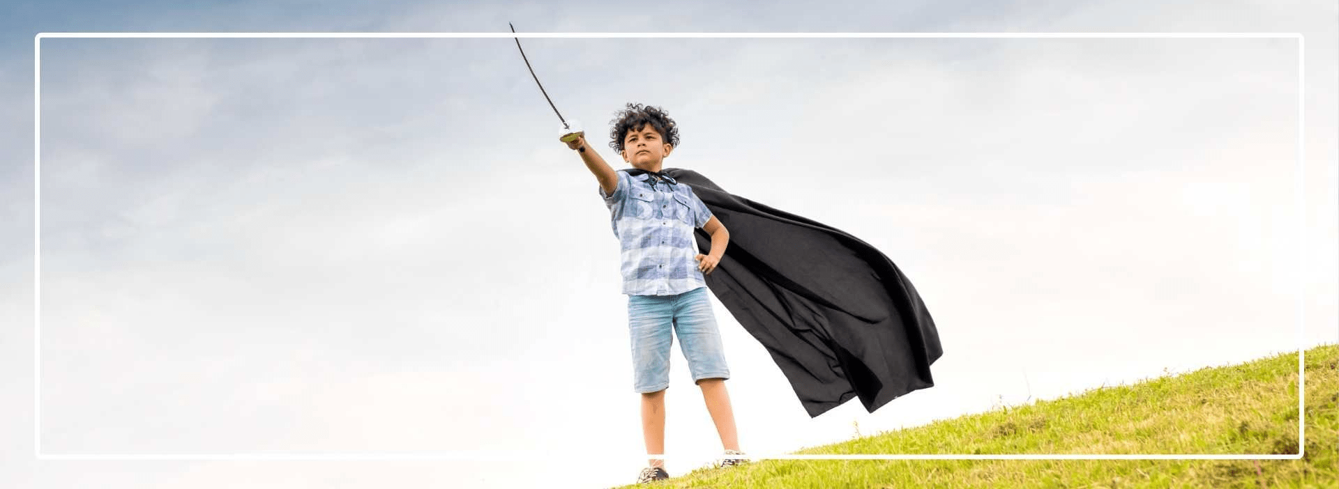 Boy with a super hero cape on standing on a hill with arm stretched out holding a sword