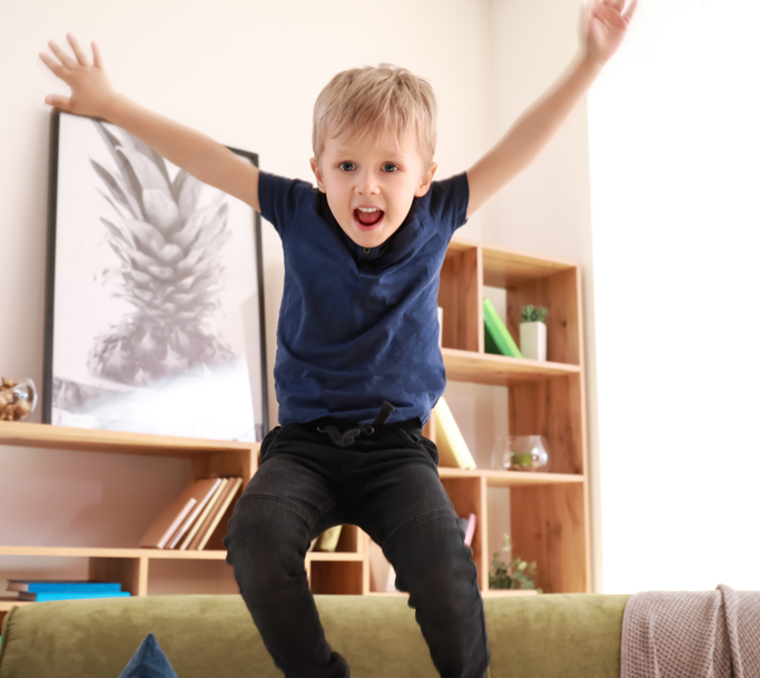 Young blonde child in a blue shirt and dark pants jumping on a green couch with arms raised high and mouth open in excitement, in a bright living room with wooden shelving