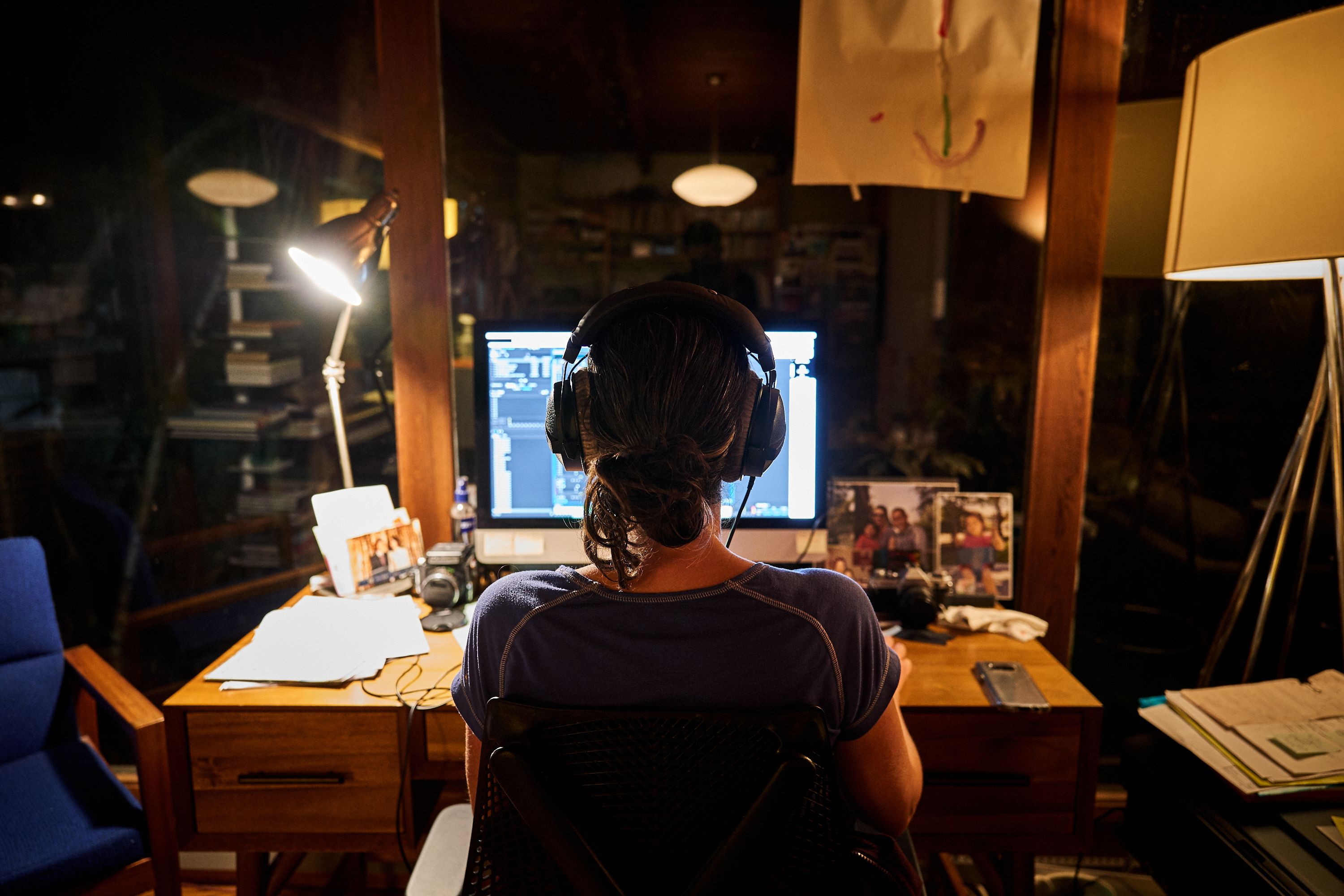 Jen sits at her desk wearing headphones, facing a computer screen in a home recording studio with a microphone, lamp, and family photos visible.